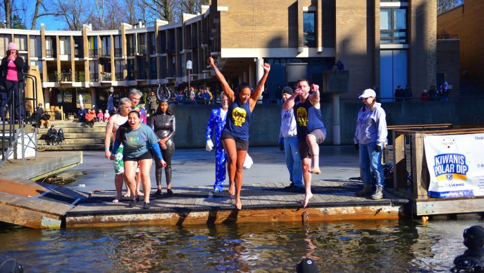Three people line up on a wooden platform to jump into a lake. The woman on the left is about to jump, the woman in the middle is mid-jump with her arms in the air, and the man on the right is mid-jump and pointing at the camera with both hands.