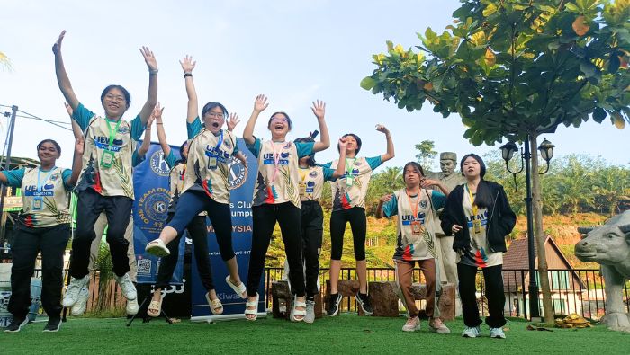 Nine Green Generation Campers jump in the air with their arms raised. Behind them are a tree, statues of a cow and a man, and a fence overlooking greenery and a house