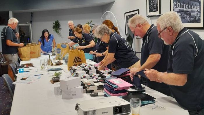 Members of the Kiwanis Club of Brisbane line up on either side of a long table, creating an assembly line to insert school supplies into pencil bags