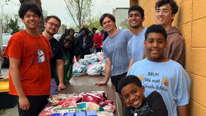 A group of volunteers poses over a long table with food packets at one end, hygiene kits at the other. Johns Hopkins University CKI's Hygiene Kits for the Homeless event, 2025