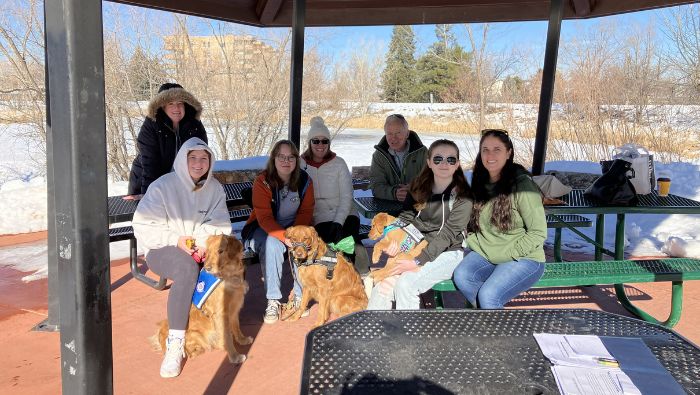 Un grupo de personas sentadas alrededor de mesas de picnic en un parque en invierno. Tres de ellas tienen golden retrievers con arneses.