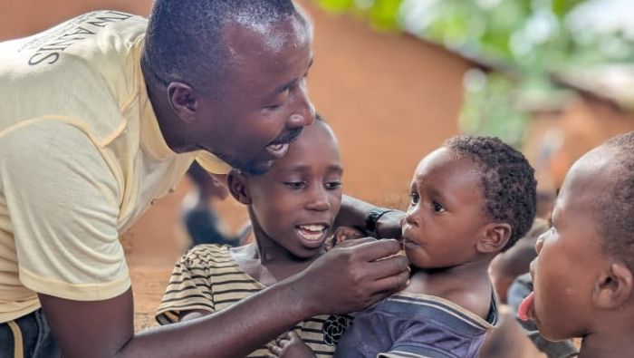A man administers medication to a toddler being held by an older child. A third child stands to the side, sticking their tongue out.